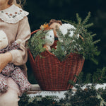 Olli Ella Christmas Dinky Dinkum dolls are in the red Poinsettia Carry Basket, and are hiding behind pine tree branches. A child is sat next to the basket which is on a white wall, and is holding a Dozy Dinkum doll in their hands