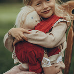 A child is sat down on a antique wooden chair, and is carrying a Dinkum Doll in the Olli Ella Dinkum Dolls Petal Carrier - Red Velvet. The child is happy and is cuddling the doll.