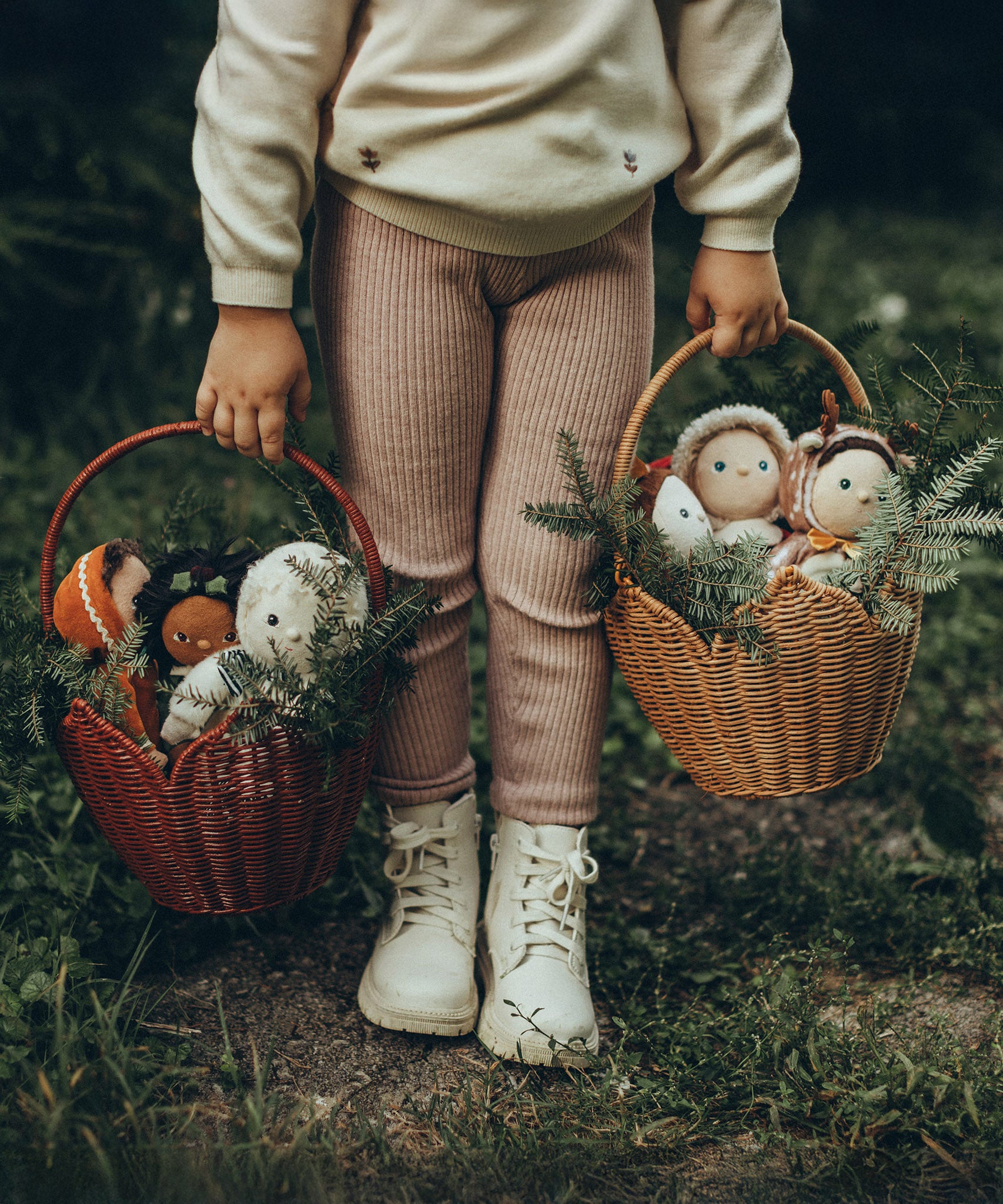 A child is holding a Natural and Red Olli Ella Rattan Poinsettia carry basket by the handles, each basket is filled with Christmas Jolly Dolly  Dinky Dinkum dolls, and the basket is decorated with pine tree branches
