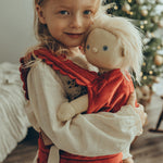 A child is carrying a Dinkum Doll in the Olli Ella Dinkum Dolls Petal Carrier - Red Velvet. The child is happy and is cuddling the doll, and there is a Christmas tree in the background