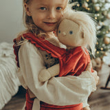 A child is carrying a Dinkum Doll in the Olli Ella Dinkum Dolls Petal Carrier - Red Velvet. The child is happy and is cuddling the doll, and there is a Christmas tree in the background