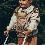 A child carrying a Dozy Dinkum Doll in a Olli Ella Dinkum Dolls Carrier in Garland print, on their front and is pushing a doll in an Olli Ella Stroller