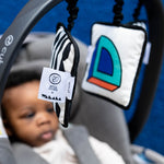 A baby sat in a car seat, looking at the Etta Loves x Camille Walala Sensory Hanging Squares which are hanging from the carry arm of the car seat
