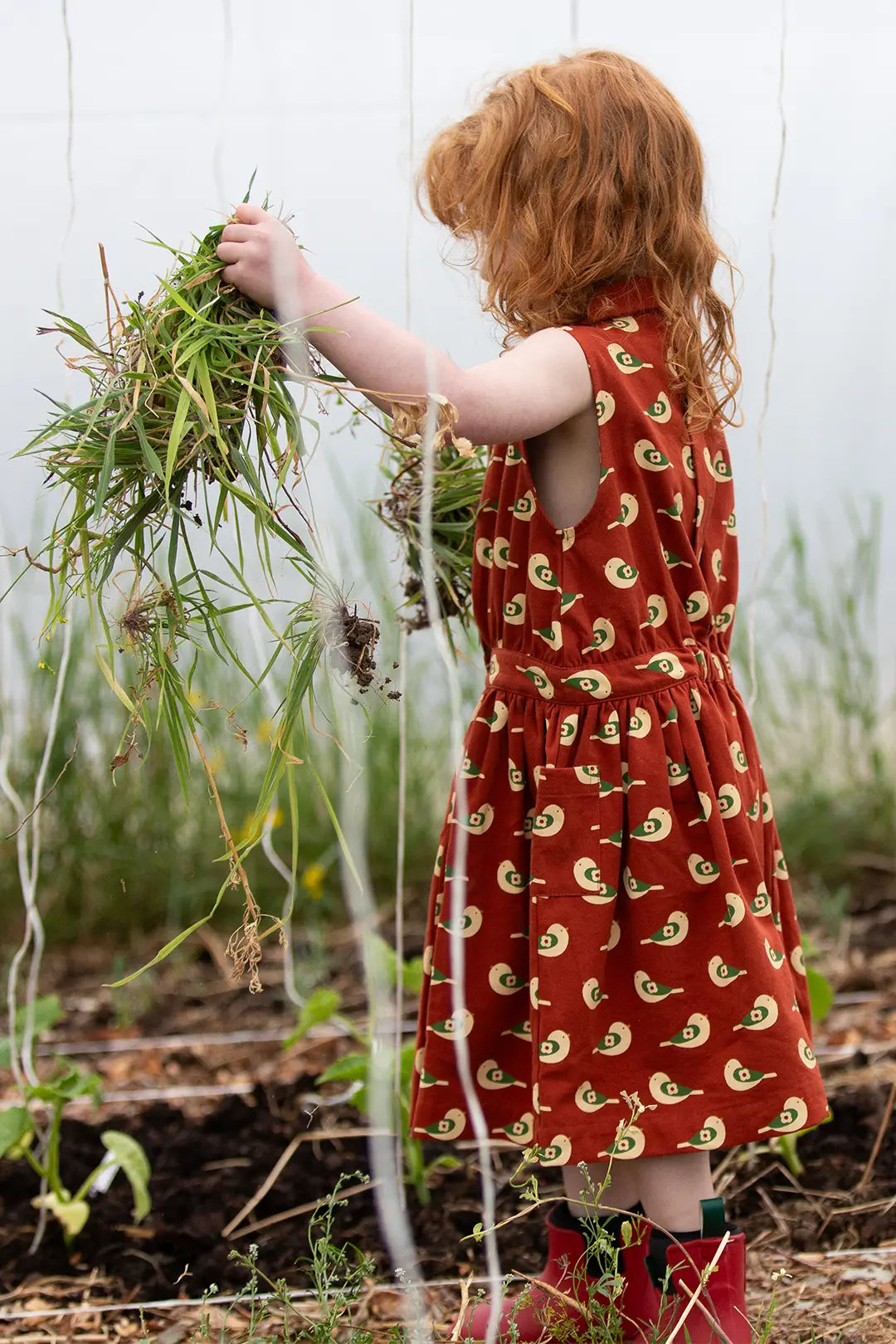 Child in red Little Green Radicals sleeveless dress and pocket with bird pattern holding plants in a garden.