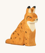 Ostheimer Wooden Sitting Lynx Toy. A beautifully handmade and crafted Wooden Lynx figure with delicate black spots, ear tips and face, with an orange body and natural wood chest. The lynx is sitting on a cream background
