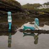 Ostheimer handmade in Germany wooden sitting Mermaid figure next to a lying mermaid figure. Handpainted in vibrant blue and resting on a rock by water and both sold at Babipur.