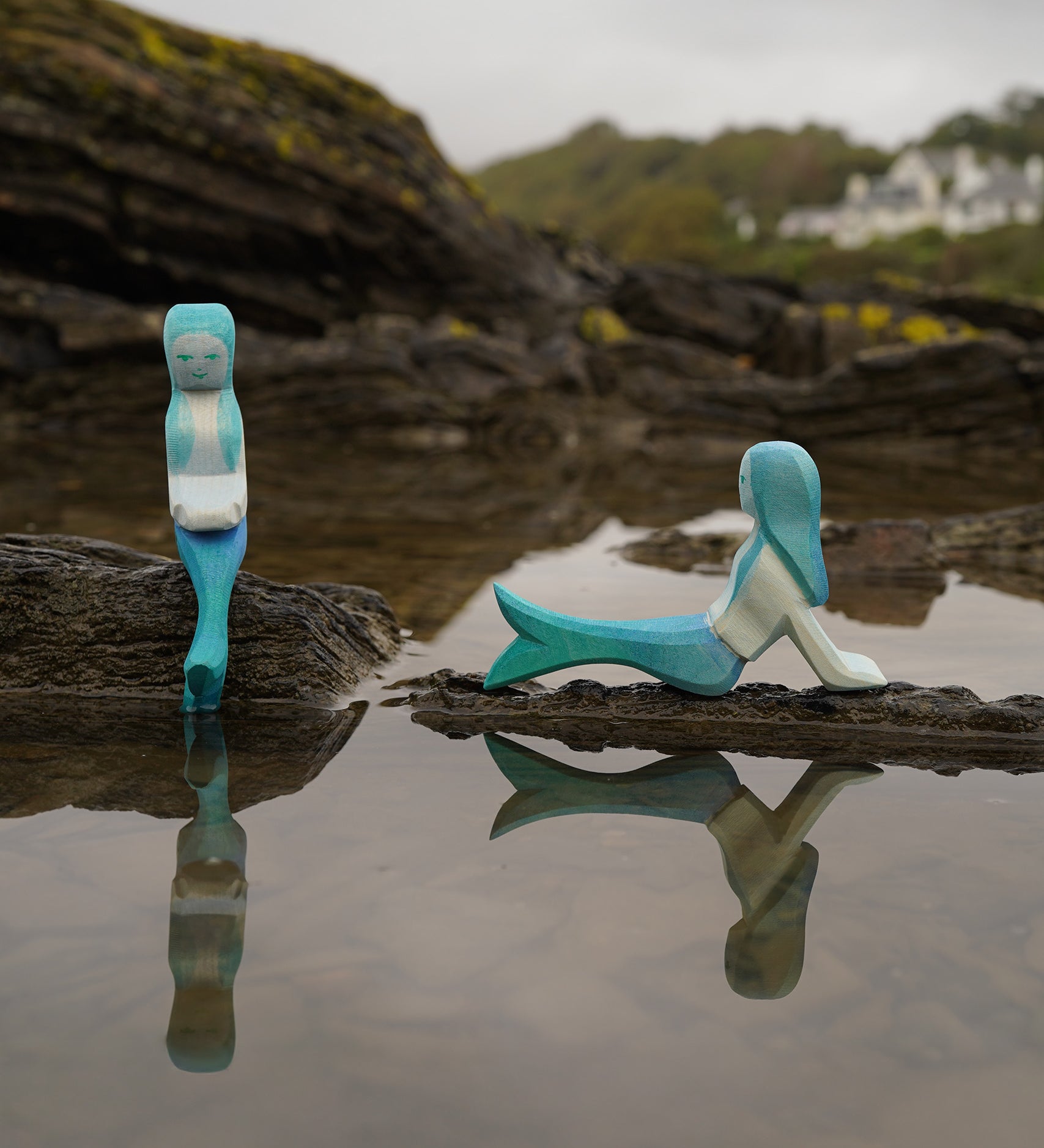 Ostheimer handmade in Germany wooden sitting Mermaid figure next to a lying mermaid figure. Handpainted in vibrant blue and resting on a rock by water and both sold at Babipur.