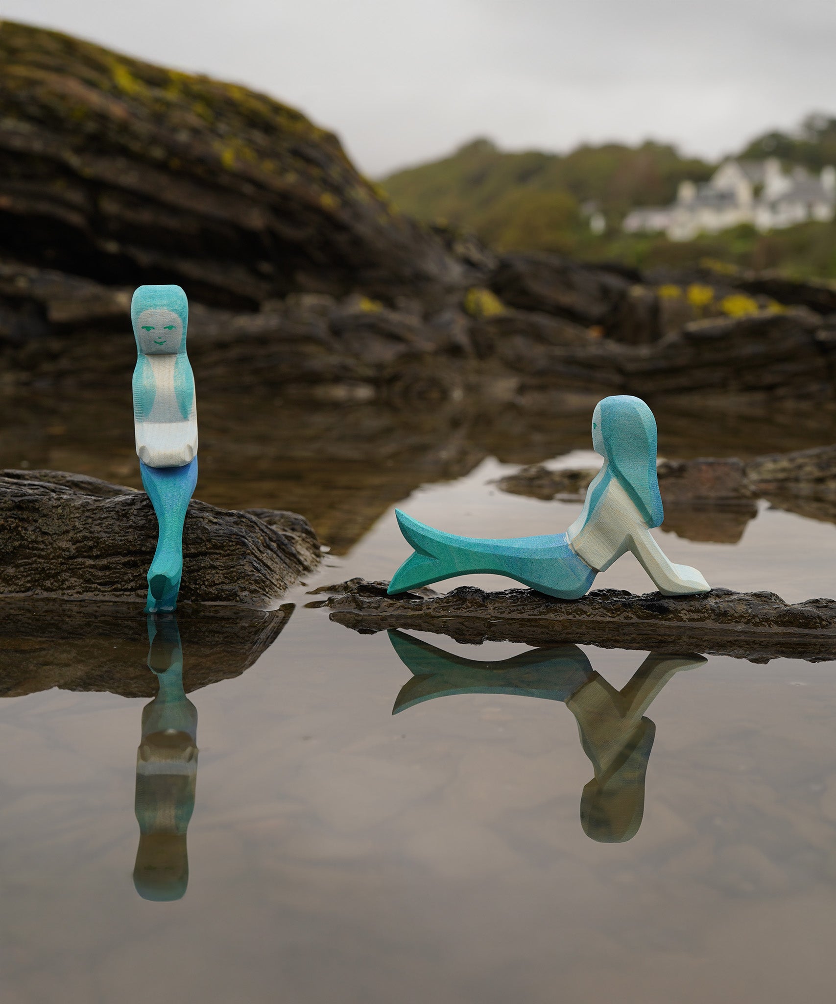 Ostheimer handmade in Germany wooden sitting Mermaid figure next to a lying mermaid figure. Handpainted in vibrant blue and resting on a rock by water and both sold at Babipur.