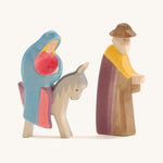 A wooden Mary Ostheimer figure wearing a red gown and blue cape sitting on a grey donkey with a Ostheimer wooden Joseph figure standing next to them on a cream background.
