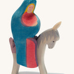 A wooden Mary Ostheimer figure wearing a red gown and blue cape sitting on a grey donkey on a cream background.