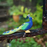 Ostheimer wooden peacock figure with it's feathers closed standing on a metal bar. Hand-painted in vibrant blues and greens, made from sustainable wood in Germany and sold at Babipur.