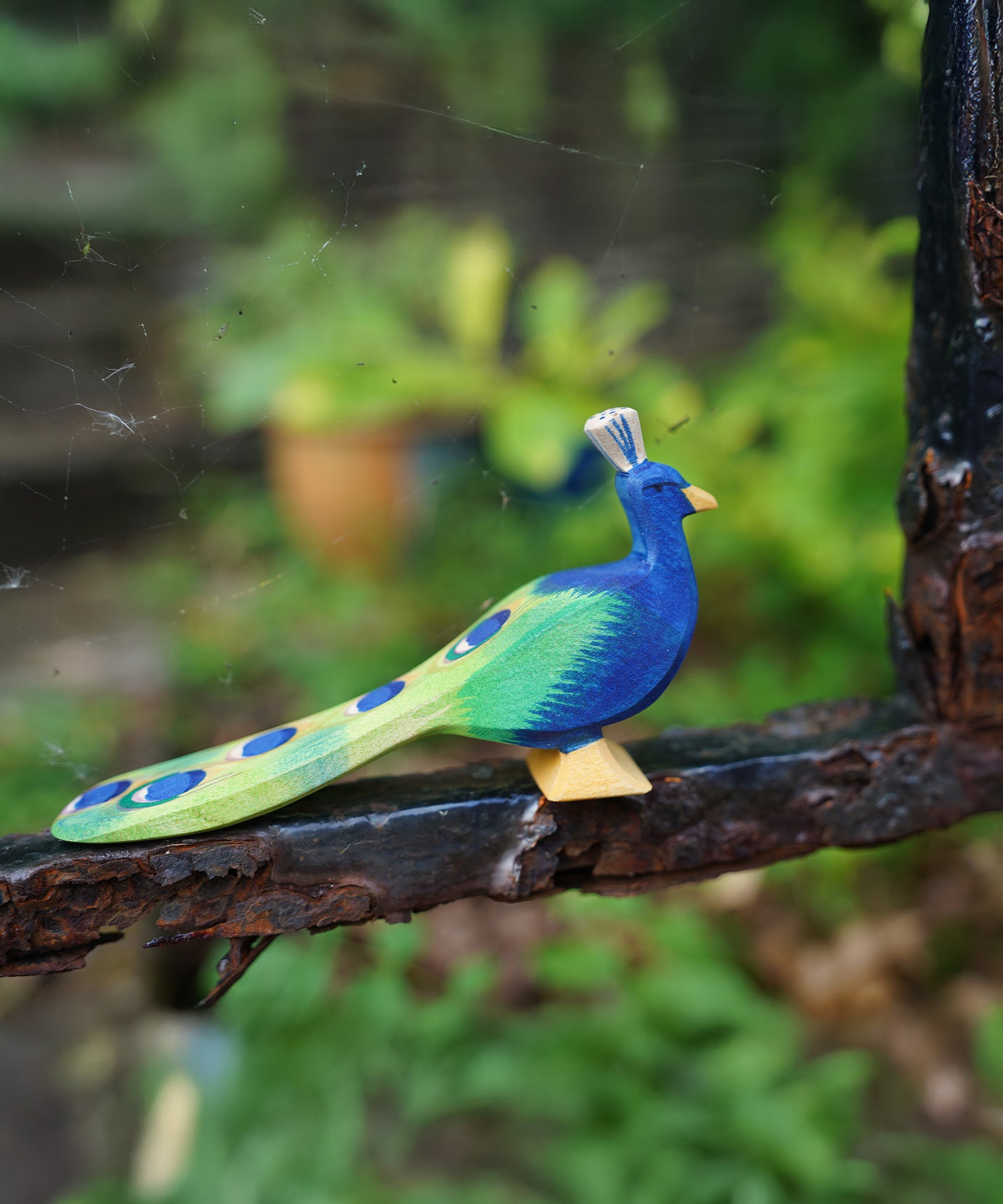 Ostheimer wooden peacock figure with it's feathers closed standing on a metal bar. Hand-painted in vibrant blues and greens, made from sustainable wood in Germany and sold at Babipur.