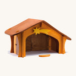 A wooden Ostheimer stable with a wooden shooting star on the top and a wooden bird perch in front of the stable on a cream background.