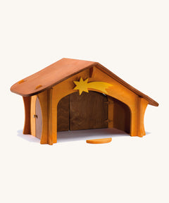 A wooden Ostheimer stable with a wooden shooting star on the top and a wooden bird perch in front of the stable on a cream background.