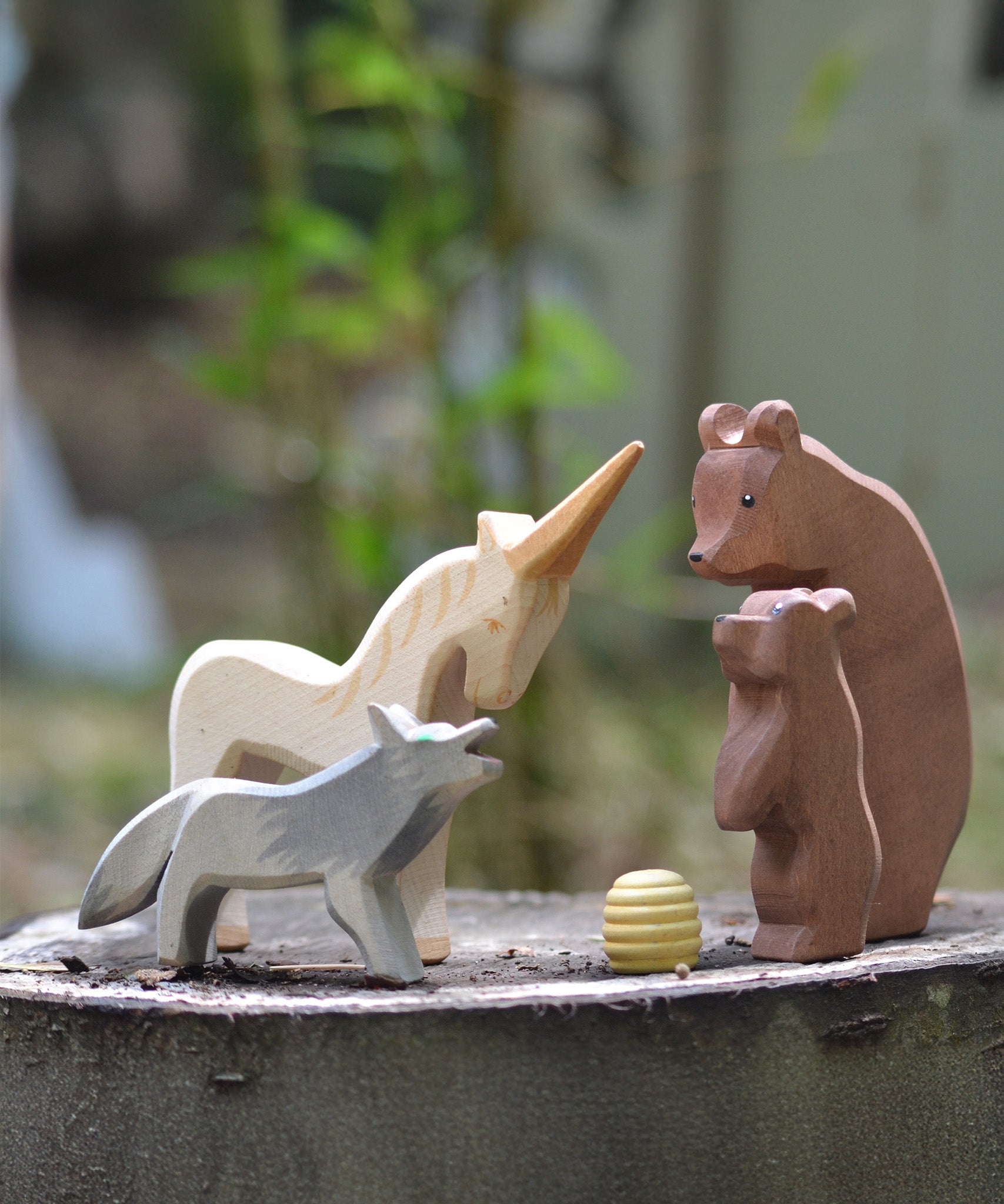 Ostheimer Bear with Cub, Howling Wolf, and Unicorn wooden figures gathered around a beehive, handmade in Germany, natural play scene on tree stump, Waldorf small world toy, sold at Babipur