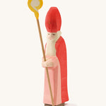  A wooden Ostheimer St Nicholas figure wearing a red gown and matching red hat holding a wooden staff on a cream background.