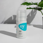 The Green People OY! Organic Teen Deodorant is stood on a white table, with a grey and white marble backgoound, and a green plant in the upper right corner
