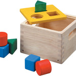 The Plan Toys Shape & Sort It Out is made with a natural wood finish with cut out handles at the sides, a yellow painted lid the square, circle, and triangle shape blocks in green, red and blue. White background. 