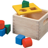 The Plan Toys Shape & Sort It Out is made with a natural wood finish with cut out handles at the sides, a yellow painted lid the square, circle, and triangle shape blocks in green, red and blue. White background. 