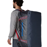 A man carrying the Patagonia black hole 100L duffel bag in smolder blue / amanita red on his back by the shoulder straps. Backpack available at Babipur. 