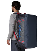 A man carrying the Patagonia black hole 100L duffel bag in smolder blue / amanita red on his back by the shoulder straps. Backpack available at Babipur. 