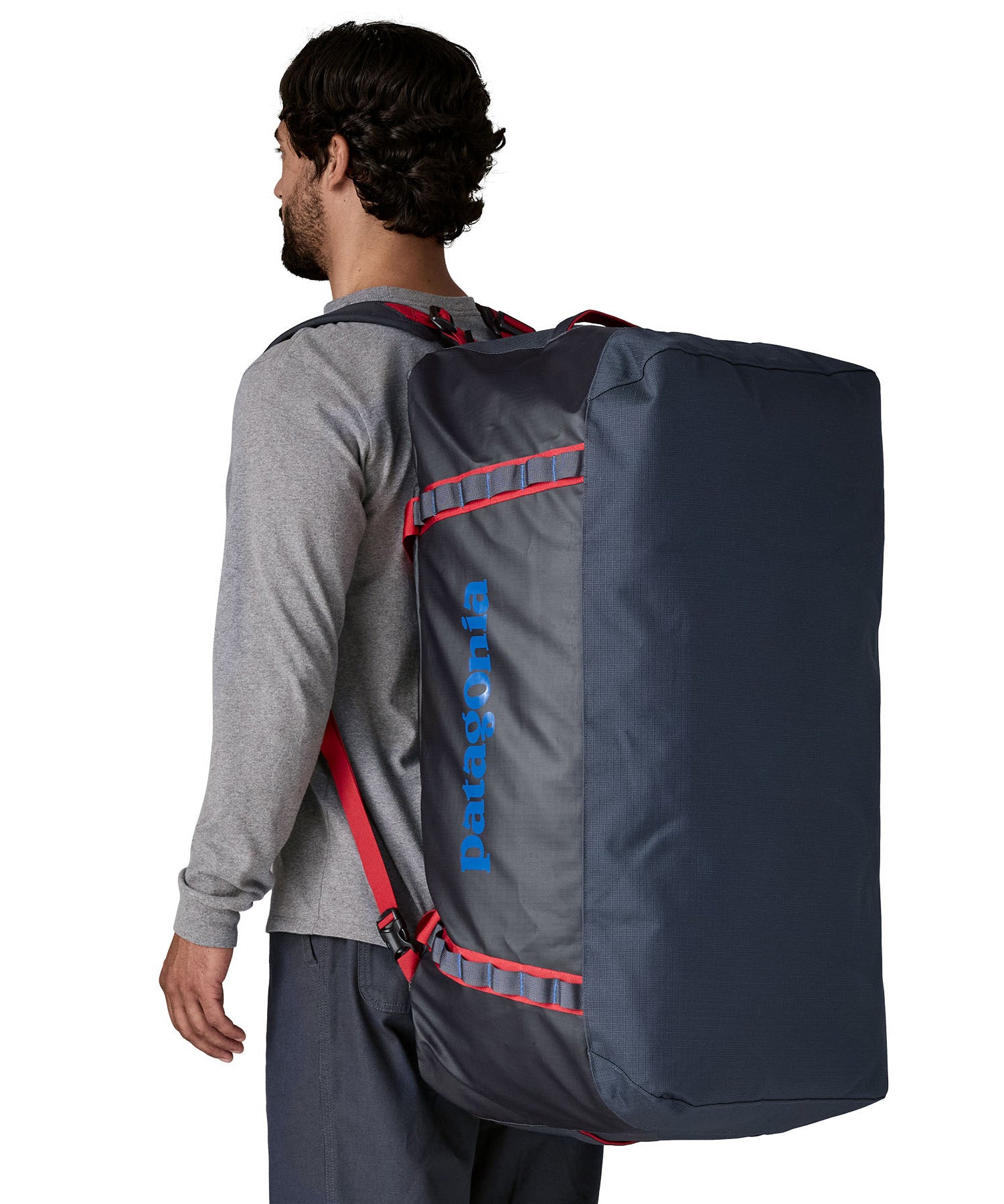 A man carrying the Patagonia black hole 100L duffel bag in smolder blue / amanita red on his back by the shoulder straps. Backpack available at Babipur. 
