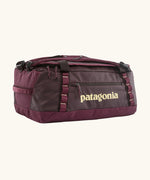 Patagonia black hole 70L duffel bag in Berry Fig. A deep maroon bag with yellow trim and Patagonia branding on the side. Features shoulders straps and a handle on the top for easy carrying.