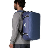 A man carrying the Patagonia black hole 40l duffel bag in current blue on his back by the shoulder straps. Backpack available at Babipur. 