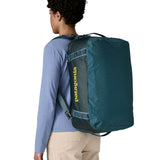 A woman carrying the Patagonia black hole 40l duffel bag in tidal teal / luminous pink on her back by the shoulder straps. Backpack available at Babipur. 