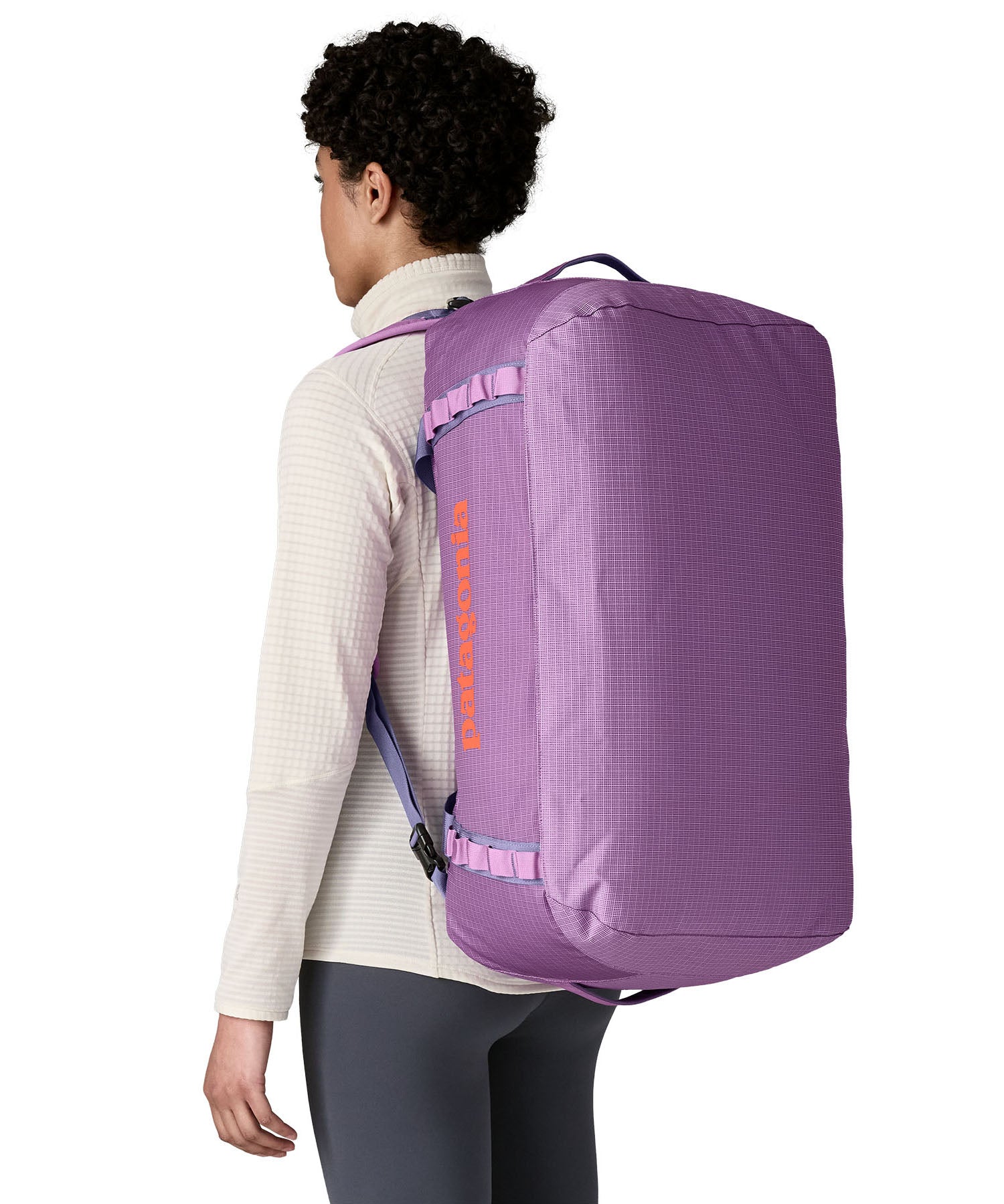 A woman carrying the Patagonia black hole 55L duffel bag in brisk purple on her back by the shoulder straps. Backpack available at Babipur. 