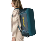 A woman carrying the Patagonia black hole 55L duffel bag in tidal teal / luminous pink on her back by the shoulder straps. Backpack available at Babipur. 