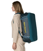 A woman carrying the Patagonia black hole 55L duffel bag in tidal teal / luminous pink on her back by the shoulder straps. Backpack available at Babipur. 