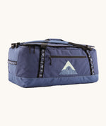 Patagonia black hole 70L duffel bag in current blue available at Babipur. A blue bag with black trim and a mountain graphic on the side. Features shoulders straps and a handle on the top for easy carrying.
