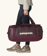 Woman carrying Patagonia black hole duffle 70L in berry fig using handle straps. 