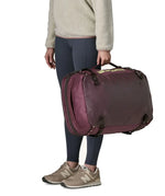 An adult carrying a Patagonia 45 litre black hole travel backpack in maroon using the carry strap on the side
