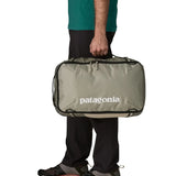 Side of man carrying a Patagonia 30 litre black hole travel backpack in beige over the shoulder using the carry handle 