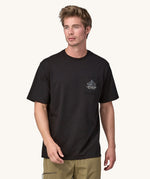 A male standing in front of a cream background. Wearing green walking trousers and the Patagonia men's organic cotton short sleeve t-shirt in black with a a small pouch pocket on the front