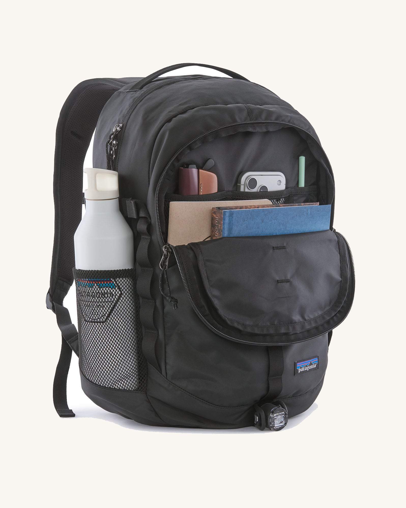 Patagonia refugio backpack in black with open front compartment with stationery placed inside and a bottle in the side holder.