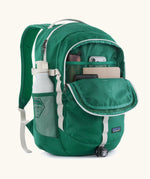 Patagonia 26L refugio backpack in green with open front compartment with stationery placed inside and a bottle in the side holder.