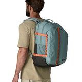 Back of man wearing Patagonia's refugio 32L daypack in sage blue with orange trim showing the mesh side pocket
