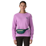 A woman wearing the Patagonia terravia mini bum bag in cascade green available at Babipur around the waist showing the zipped compartment on the front panel