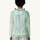 The back of the female standing in front of a cream background and wearing patterned shorts and the Patagonia lightweight jacket in light turquoise with the hood down