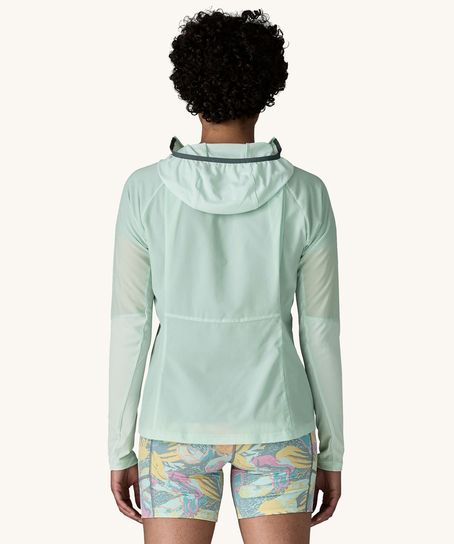 The back of the female standing in front of a cream background and wearing patterned shorts and the Patagonia lightweight jacket in light turquoise with the hood down