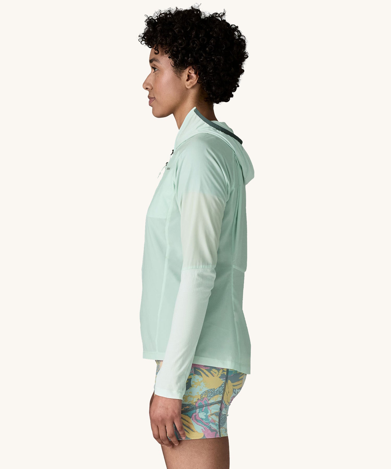 The side side of the female standing in front of a cream background and wearing patterned shorts and the Patagonia lightweight jacket in light turquoise with the hood down