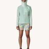 A female standing in front of a cream background and wearing patterned shorts and the Patagonia lightweight jacket in light turquoise with the hood up and zip fully fastened