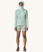 A female standing in front of a cream background and wearing patterned shorts and the Patagonia lightweight jacket in light turquoise with the hood up and zip fully fastened