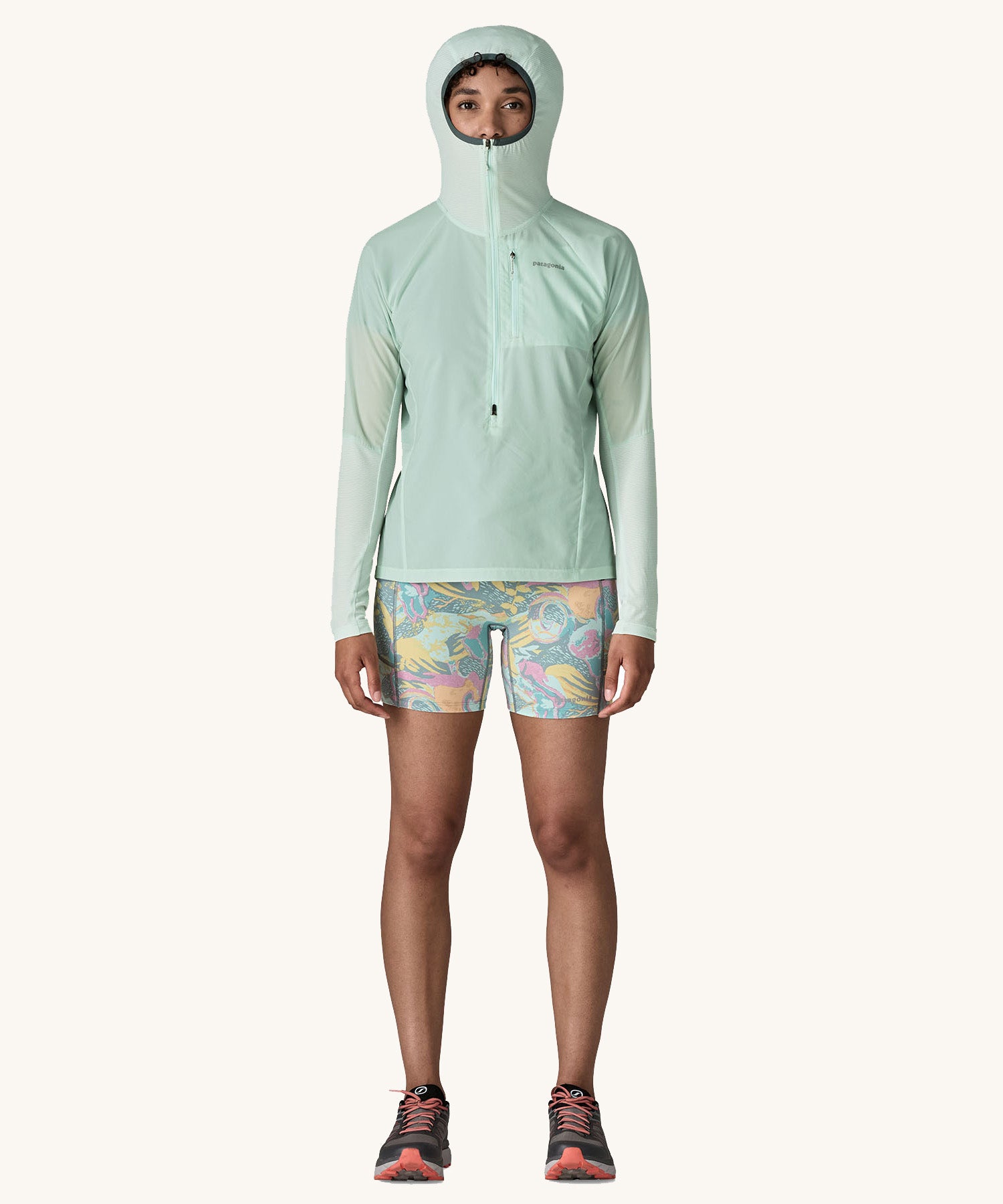 A female standing in front of a cream background and wearing patterned shorts and the Patagonia lightweight jacket in light turquoise with the hood up and zip fully fastened