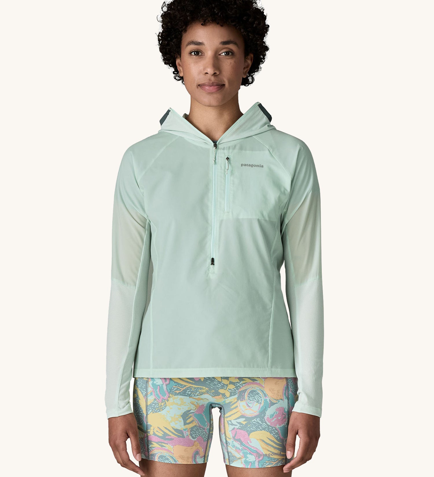 A female standing in front of a cream background and wearing floral shorts and the Patagonia lightweight jacket in light turquoise with the hood down