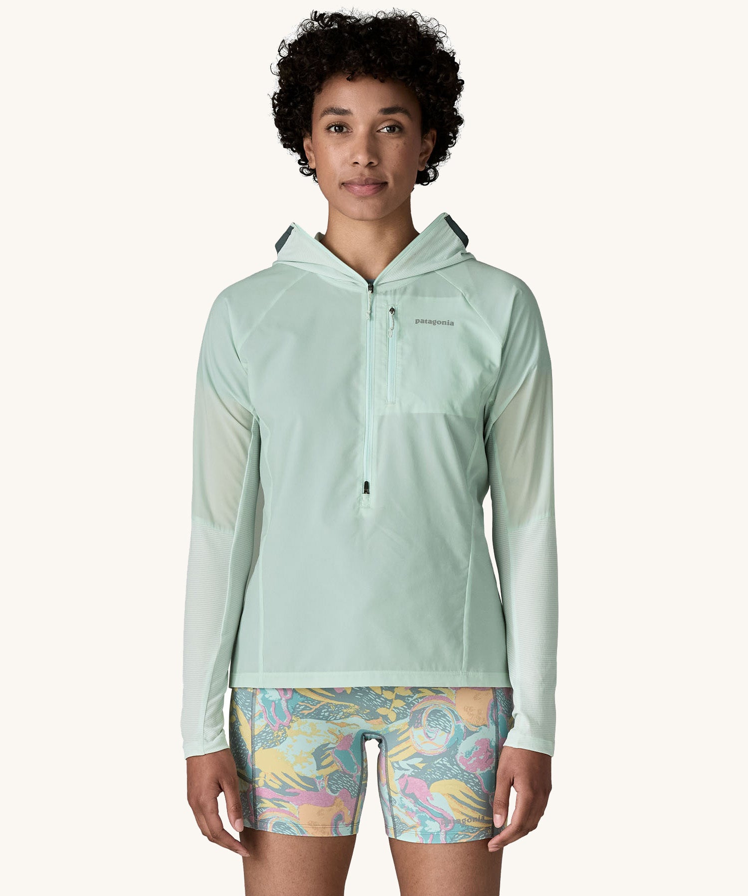 A female standing in front of a cream background and wearing floral shorts and the Patagonia lightweight jacket in light turquoise with the hood down
