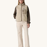 A female standing in front of a cream background and wearing a brown jumper and light cream trousers with the Patagonia thick cream fleece gilet with the zip fully fastened and has a hand in the side pocket
